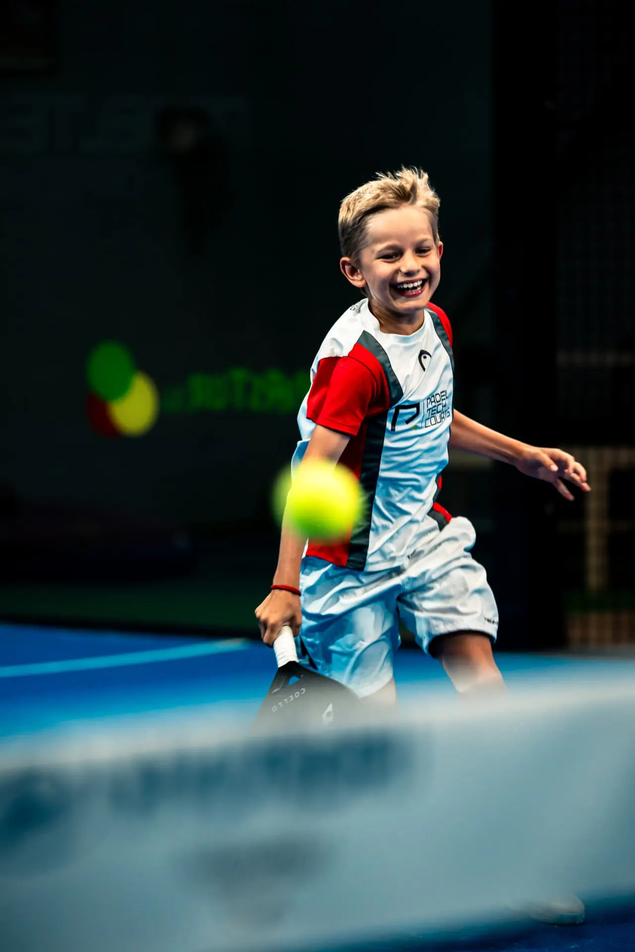Niño sonriendo mientras golpea la bola en una clase de pádel